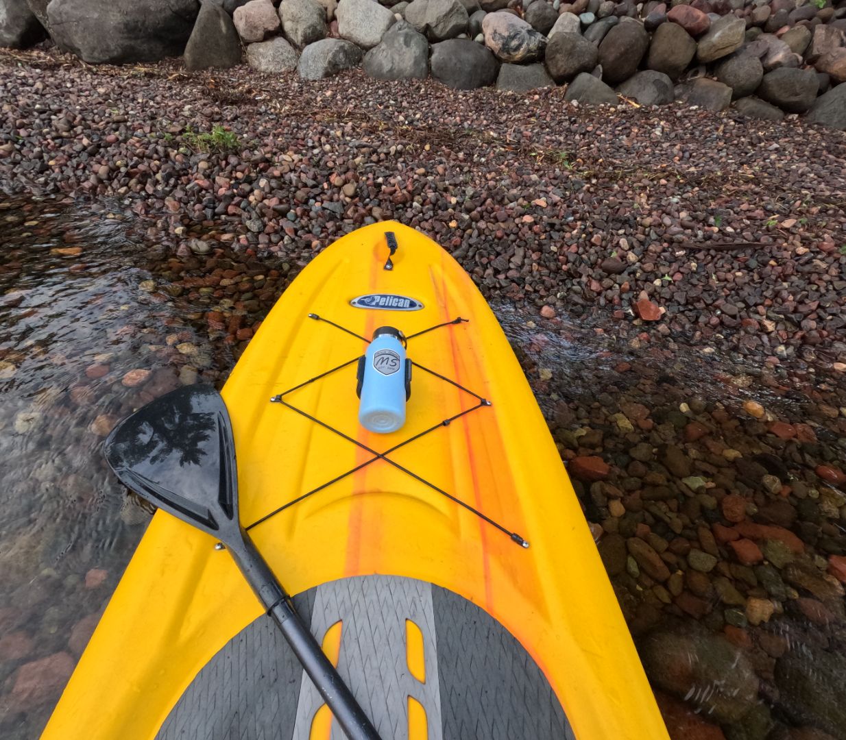 Water bottle holder on a yellow paddle board, secured with high-strength 3M VHB tape, featuring Pelican logo and paddle on rocky shore.