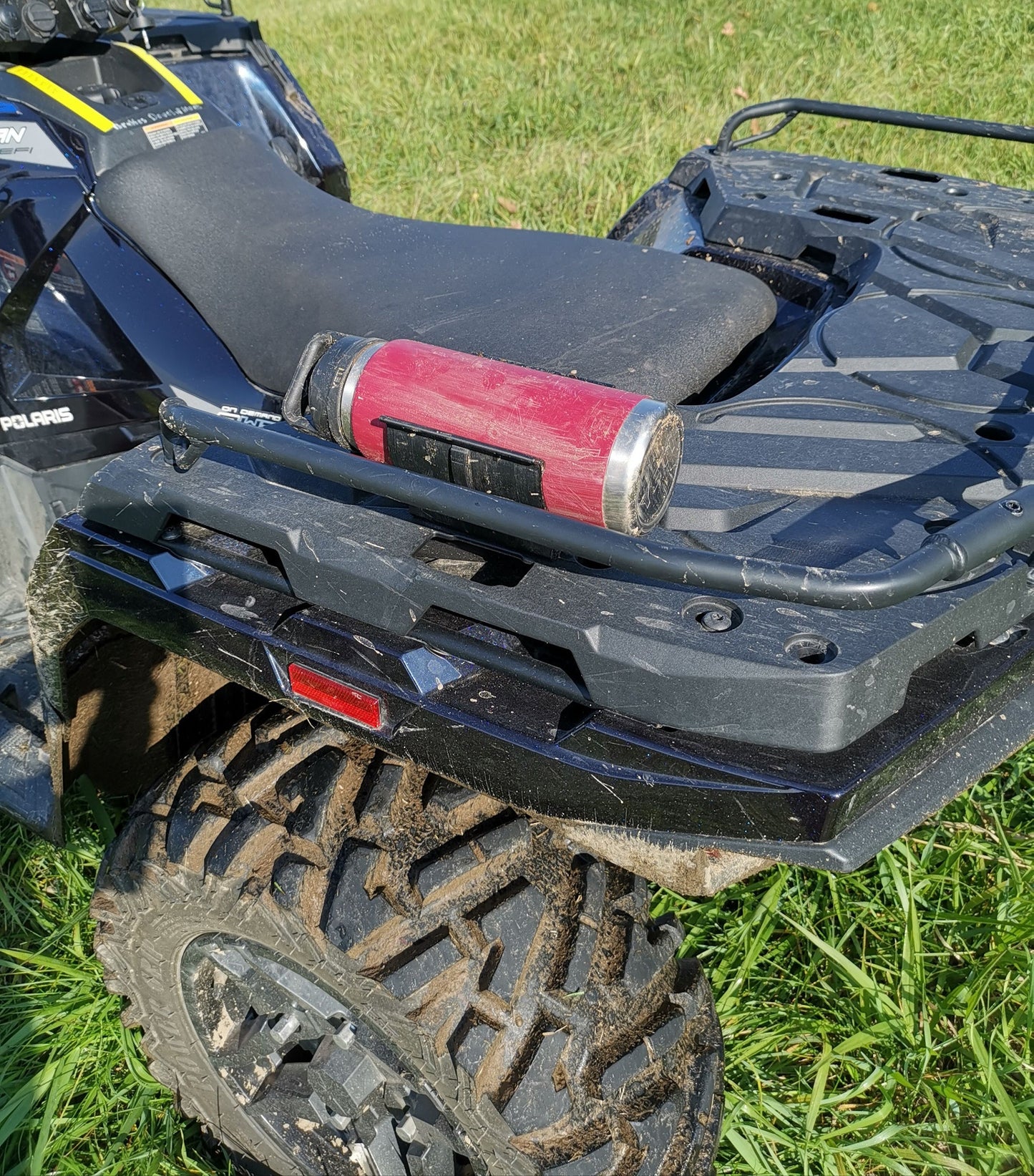 A universal holder mounted on the back of an ATV using two screws, securely holding a red water bottle, with the ATV parked on grass.