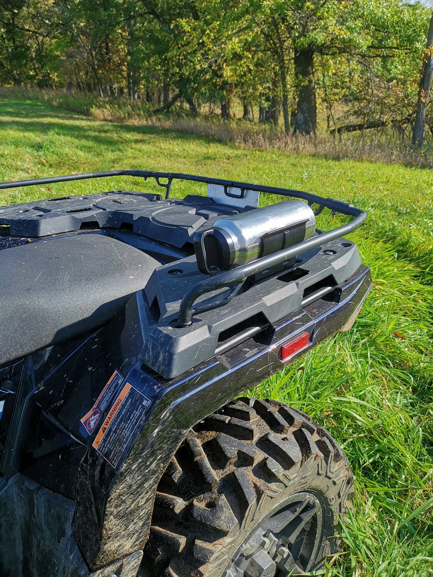 A universal holder mounted on the rear rack of an ATV, secured with two screws, holding a stainless steel thermos bottle in a grassy area.