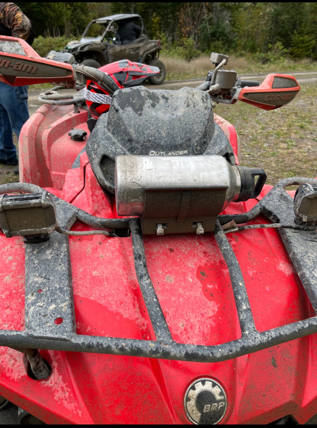 A red Can-Am Outlander ATV with a universal holder mounted on the front using two screws. The ATV is covered in mud, indicating recent off-road use.