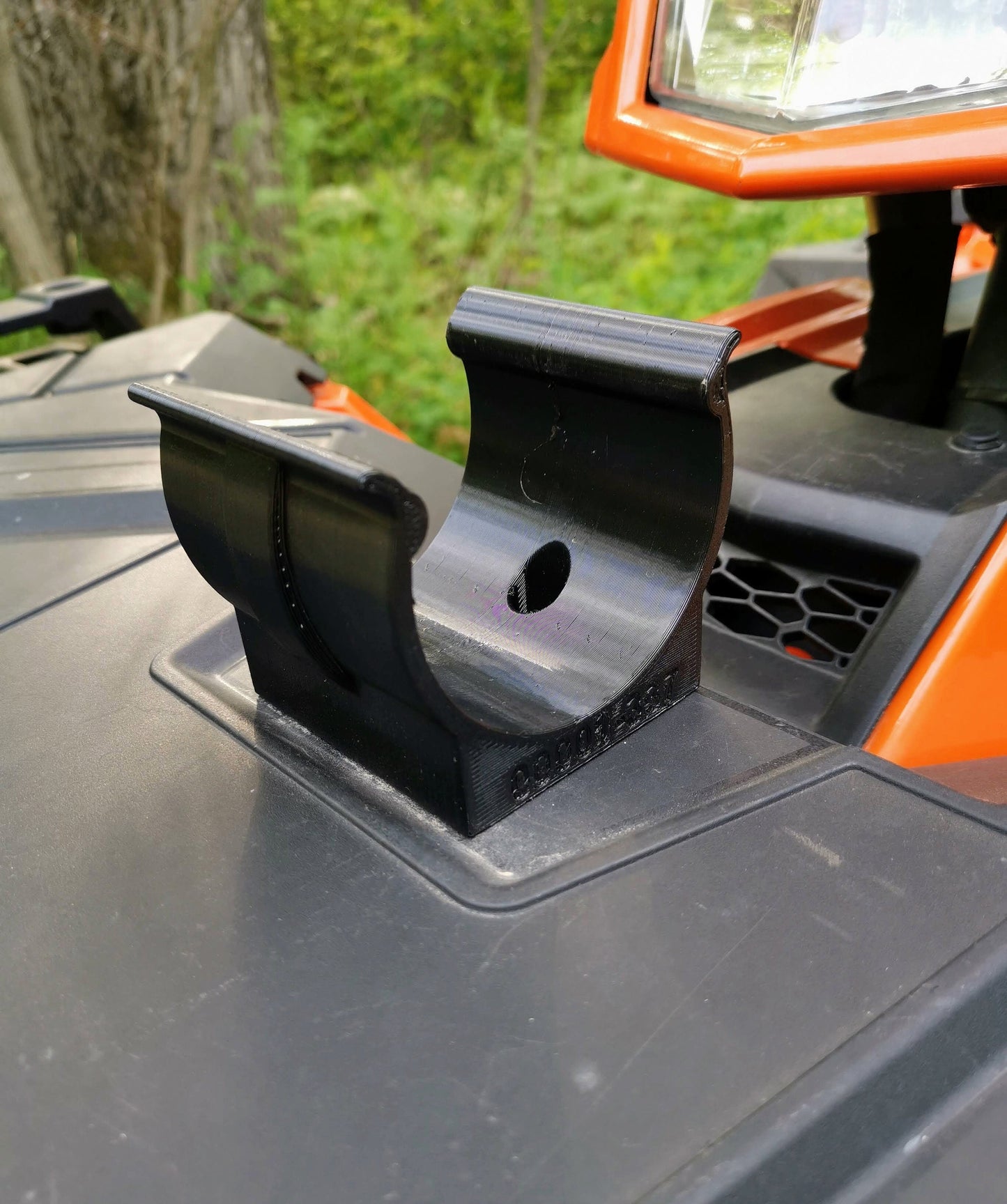 A black plastic holder mounted on the front of an orange ATV using two screws, with a forest background.