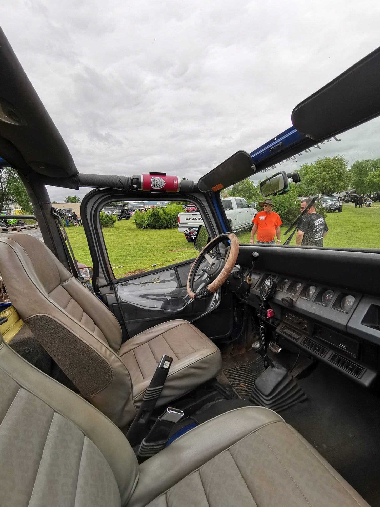 Jeep YJ interior featuring a roll bar-mounted water bottle holder, with a scenic outdoor event visible through the open door.