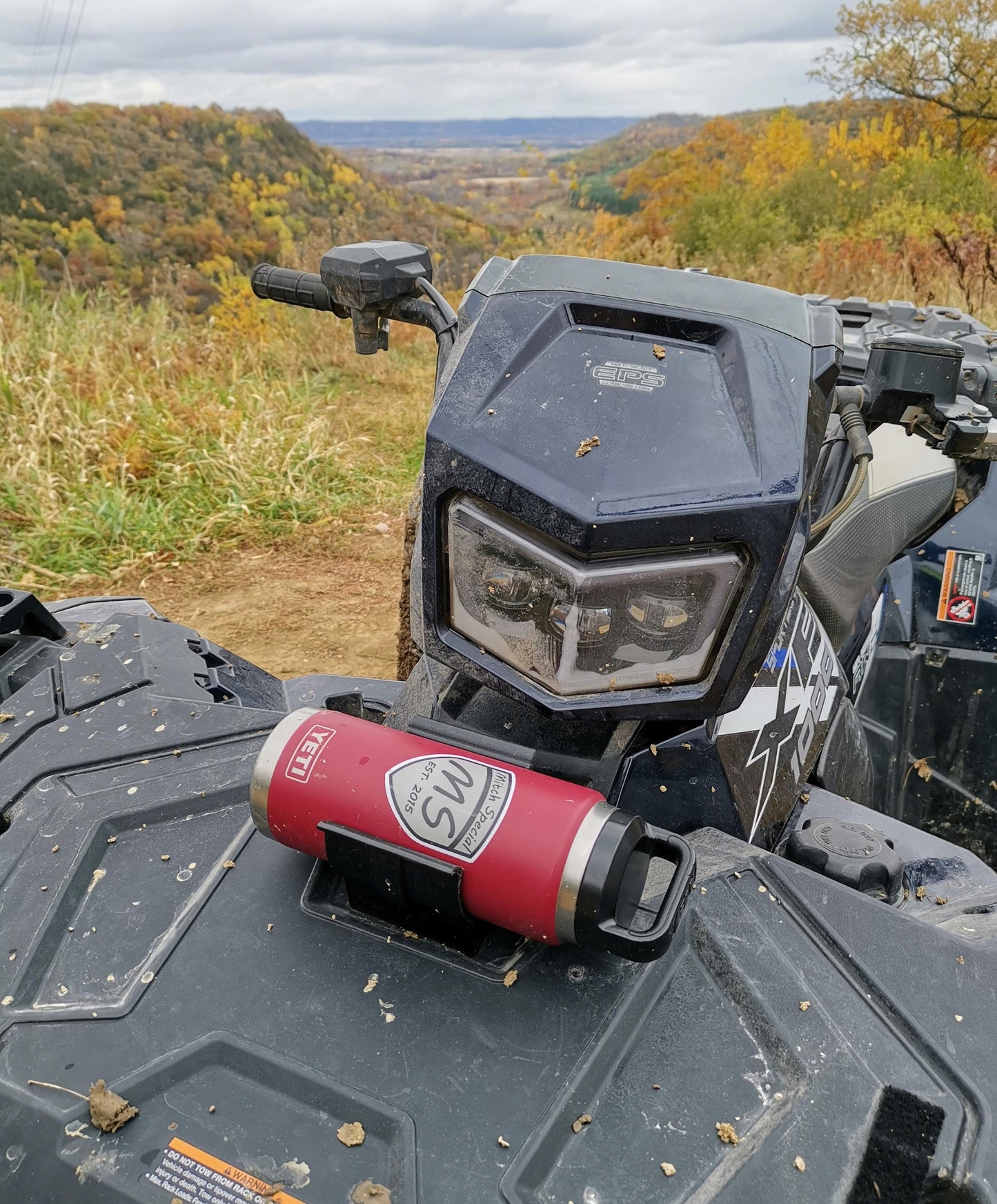 A universal holder mounted with two screws on an ATV, holding a red YETI tumbler, with a scenic autumn landscape in the background.