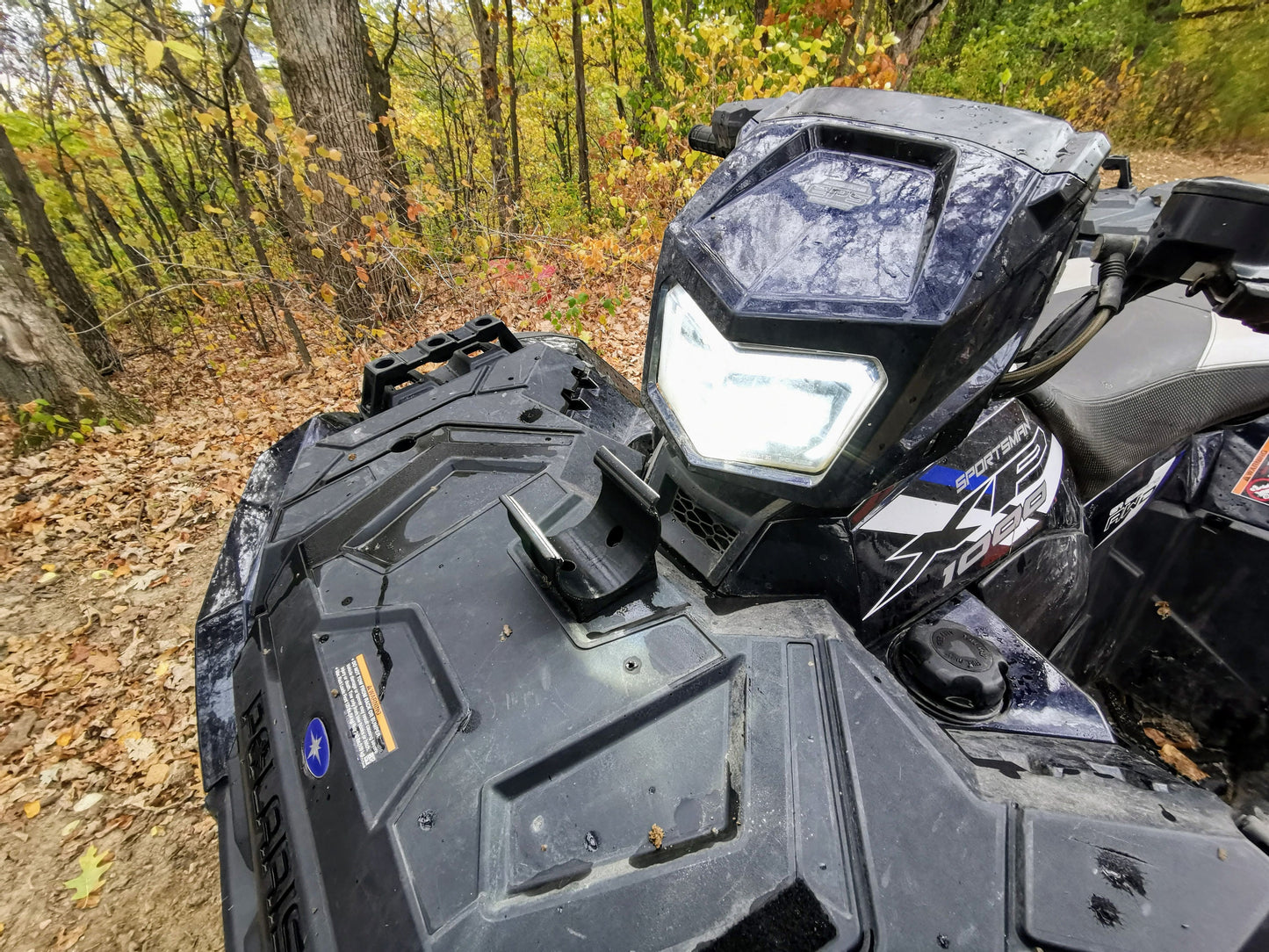 Close-up of a black ATV with a universal holder mounted on the front using two screws, surrounded by a forest with autumn leaves.