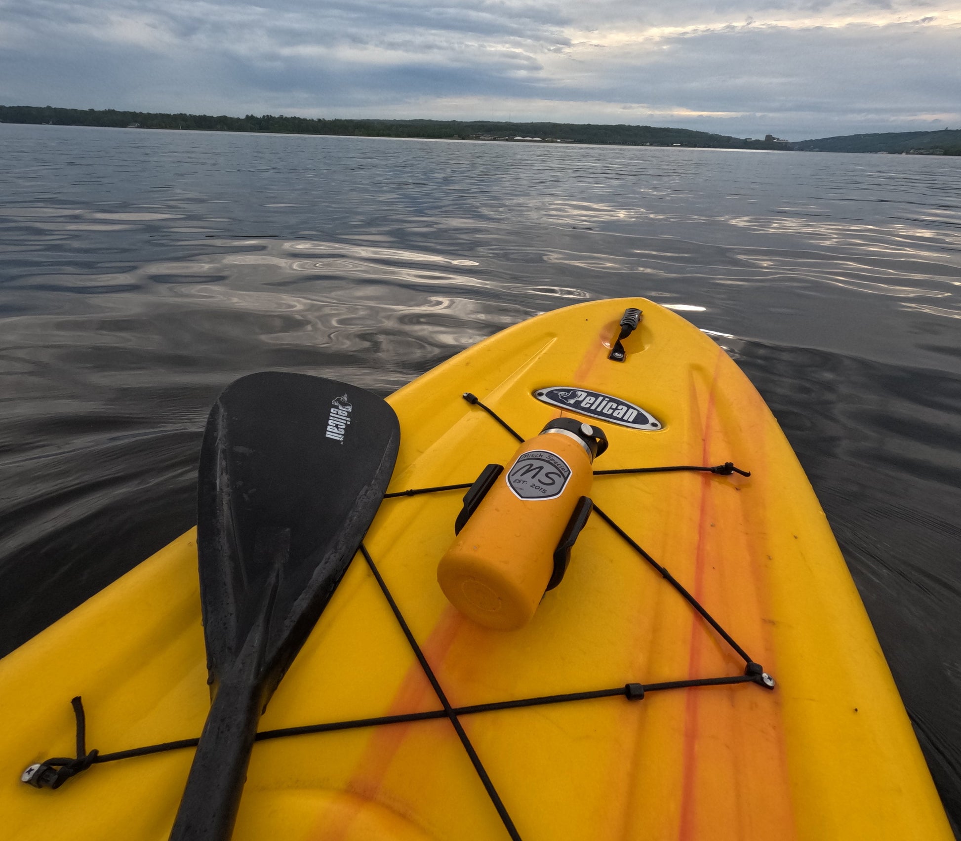 An orange water bottle with a black holder, universally mounted on a yellow kayak using 3M VHB tape, on a calm lake.