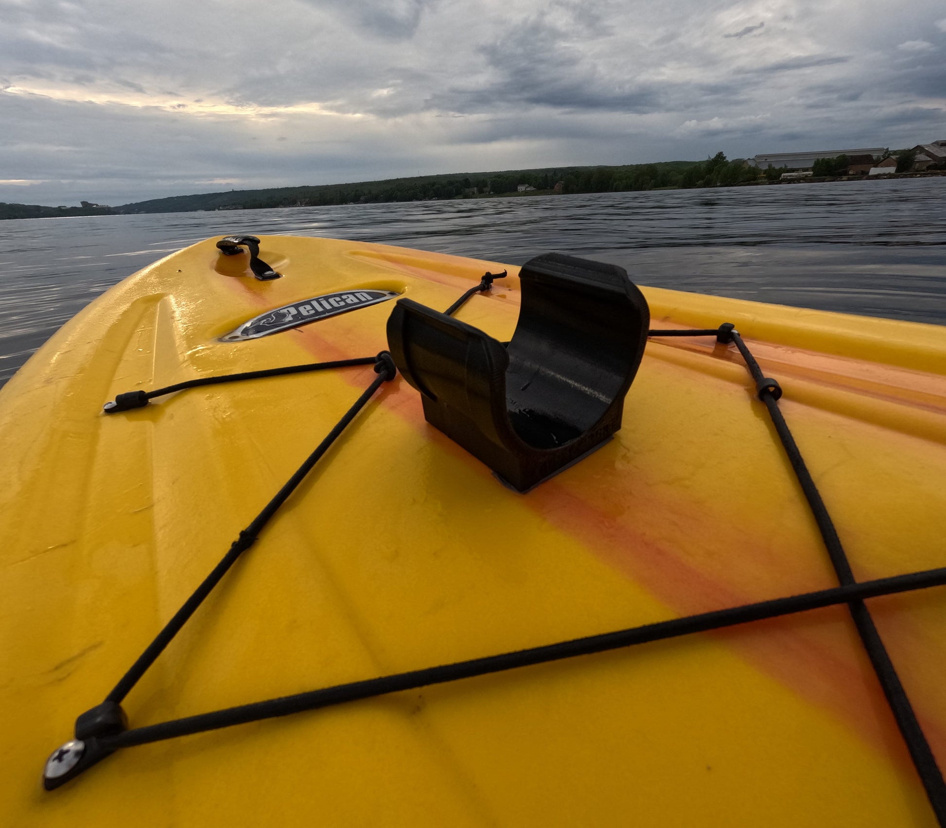 Close-up of a yellow kayak with a black universal holder mounted using 3M VHB tape, featuring a Pelican logo and bungee cords on a calm lake.