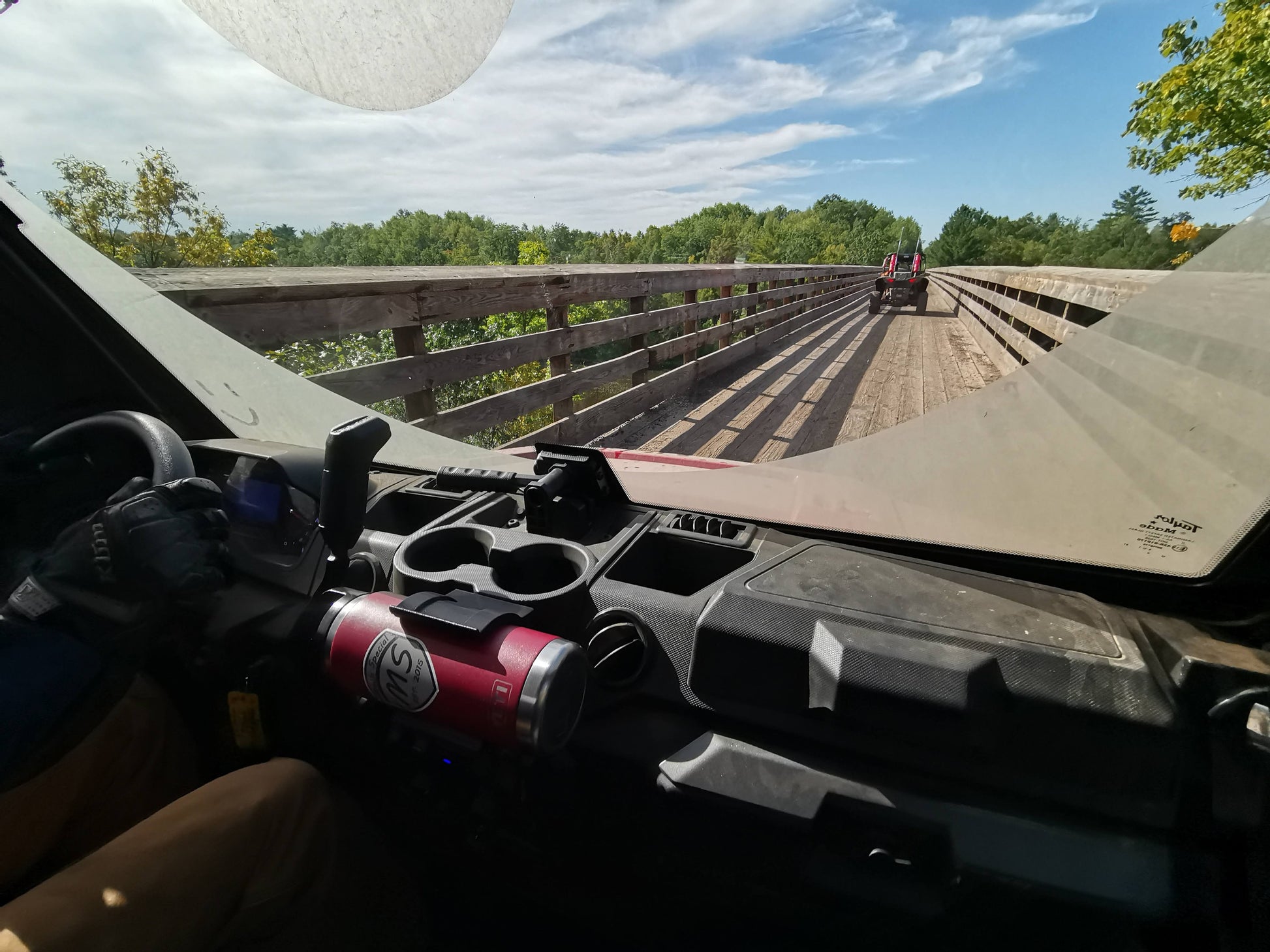 Interior view of a vehicle with a can holder mounted on the dashboard using 3M VHB tape, driving on a wooden bridge with another vehicle ahead.