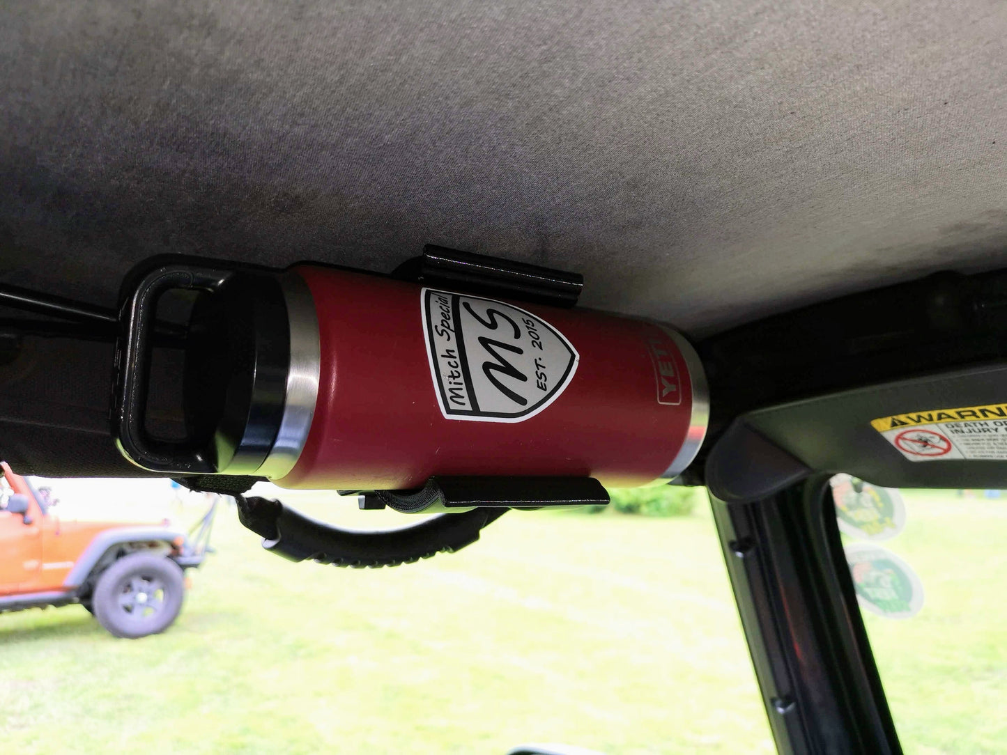 Red insulated water bottle with a 'Mitch Special' logo mounted on an overhead grab bar inside a Jeep Wrangler, showcasing a versatile storage solution for off-road and outdoor adventures.