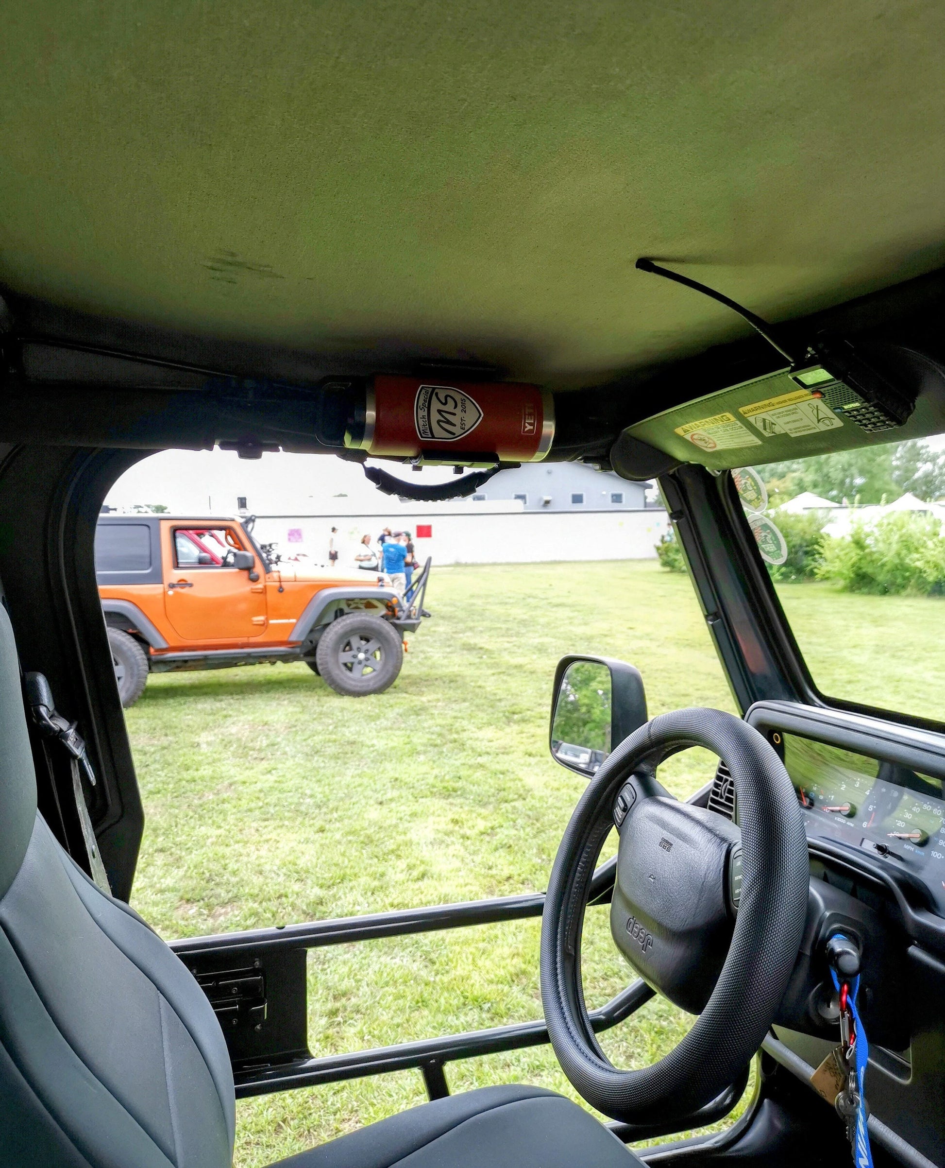 Interior view of a Jeep Wrangler featuring a red water bottle with a 'Mitch Special' logo mounted on an overhead grab bar, with an orange Jeep visible in the background at an outdoor event.