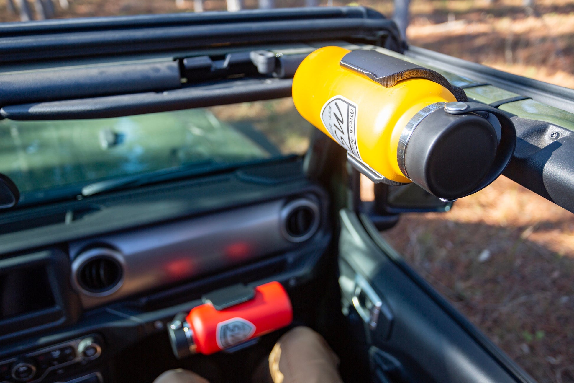 Overhead view of Jeep JL interior with tops removed, showcasing dual mounting options: water bottle holder attached to front roll bar and grab handle holder, with a passenger seated in the seat.