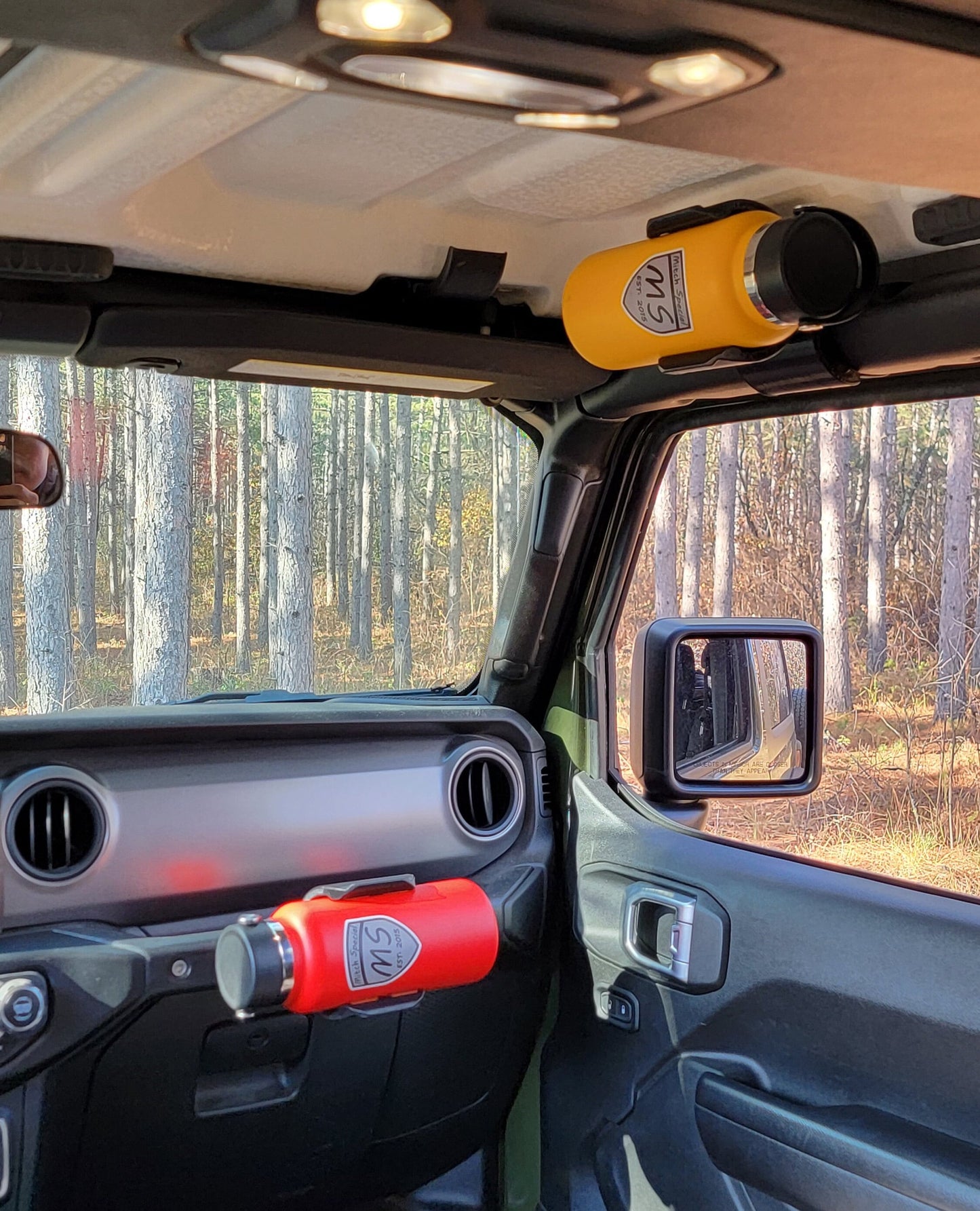 Image of Jeep JL interior, viewed from rear seat, looking over center console, showcasing dual water bottle holders: one mounted to front roll bar and another to grab handle, providing convenient hydration access for all in the Jeep. 