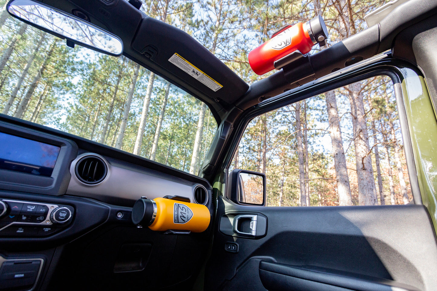 Center console perspective of Jeep JL cabin, highlighting empty water bottle holder mounted to grab handle for easy driver access, and secondary water bottle holder on roll cage providing additional storage for passengers, demonstrating versatile hydration solutions