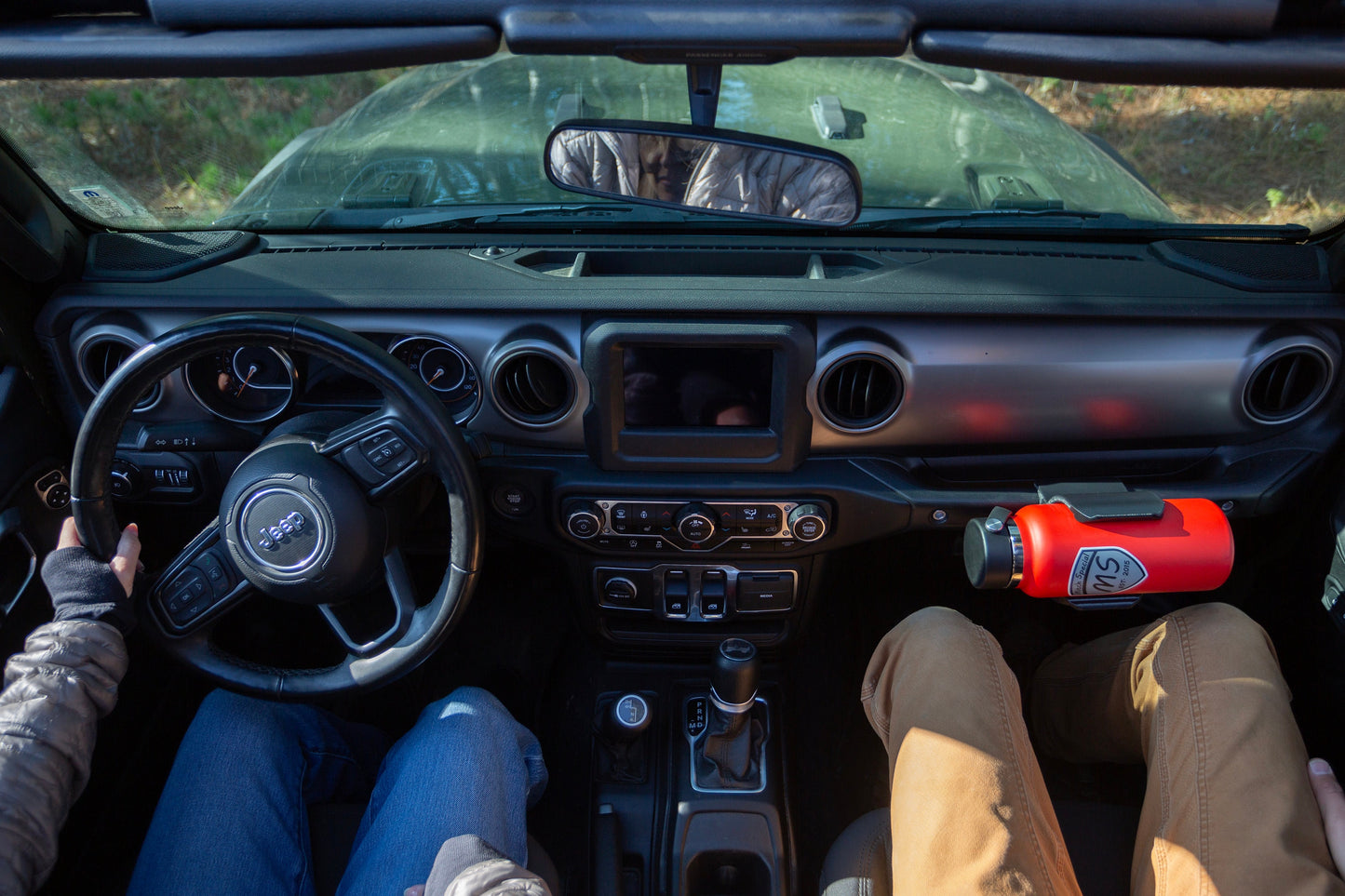 Bird's eye view of Jeep JL cabin with driver and passenger, demonstrating spacious interior and comfortable seating, with Heavy-Duty Water Bottle Holder mounted to grab handle, providing convenient hydration access without compromising legroom