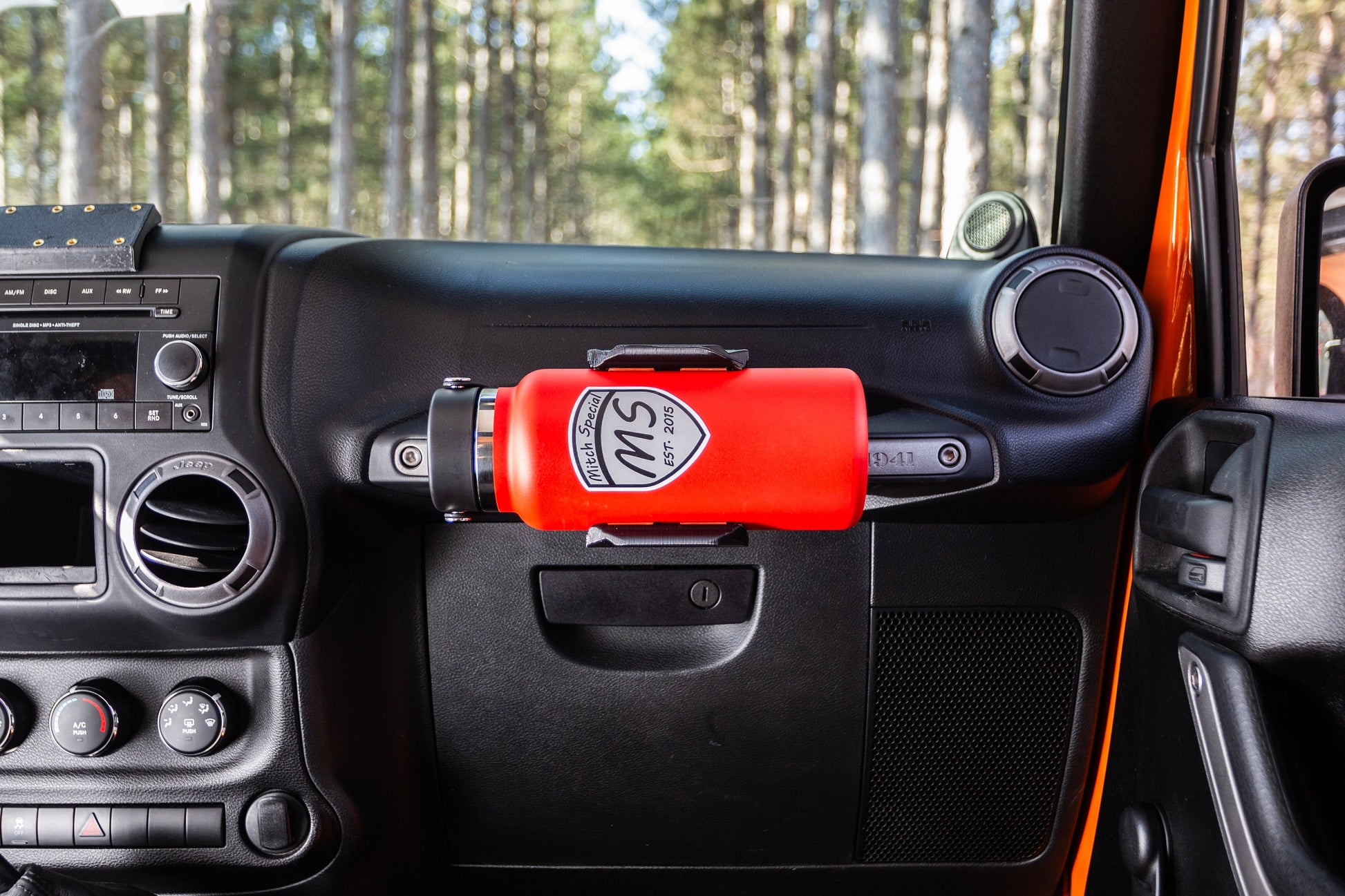 Red water bottle secured in a durable grab handle-mounted holder on the dashboard of a Jeep Wrangler, showcasing a convenient accessory for off-road adventures. Ideal for keeping beverages accessible while driving.