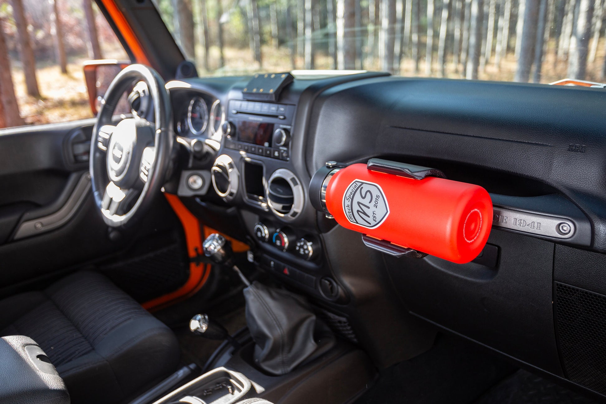 Jeep Wrangler interior with a durable water bottle holder mounted on the passenger grab handle, securely holding a red insulated bottle. Ideal accessory for off-road enthusiasts to keep hydration handy and safe while driving.