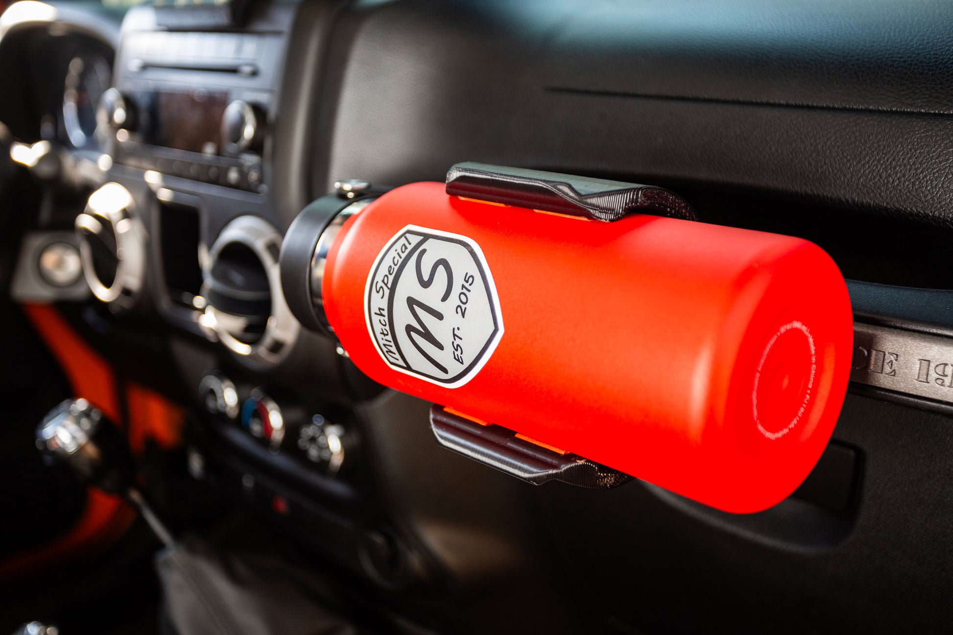 Red water bottle mounted on a Jeep Wrangler grab handle using a custom HD edition water bottle holder, designed for secure and convenient storage, with a modern car interior in the background.