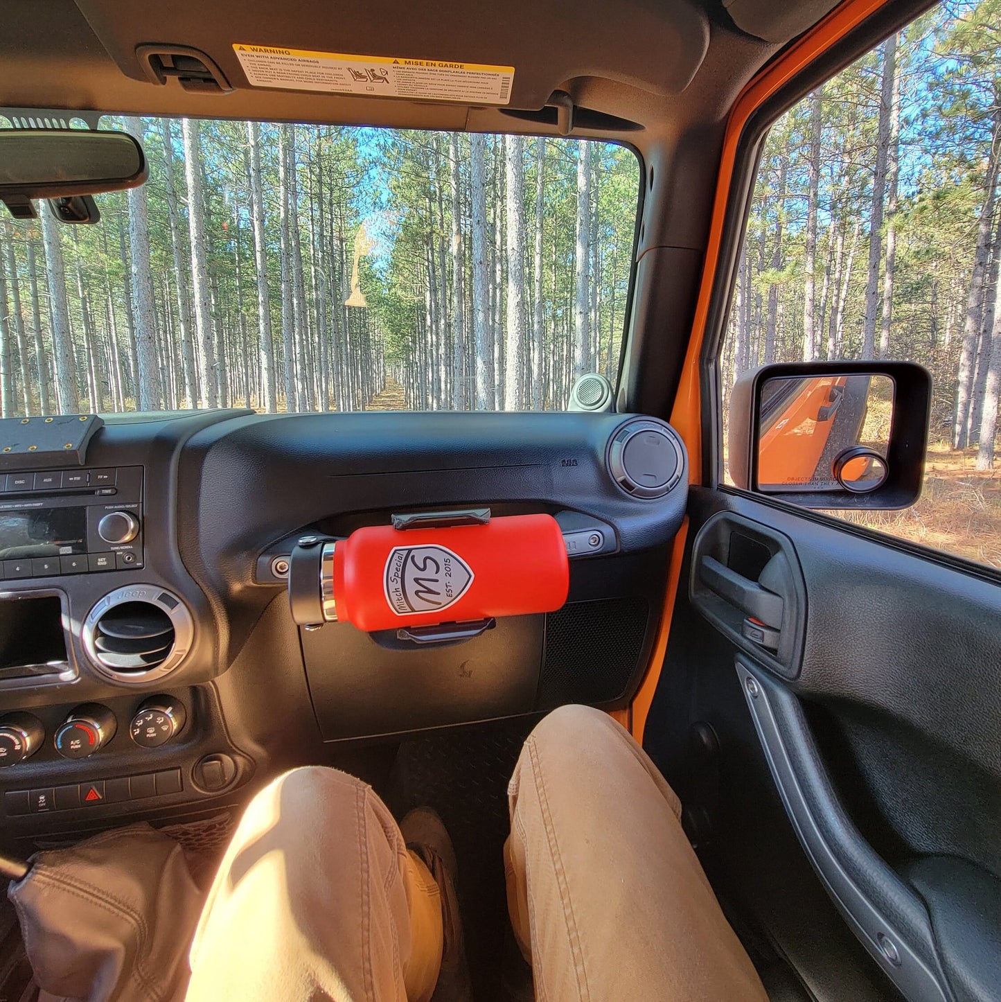 View of a Jeep Wrangler showcasing a red 'Mitch Special' water bottle securely mounted to the grab handle. Surrounded by a scenic forest backdrop, this setup highlights a practical and stylish hydration solution for outdoor and off-road enthusiasts.