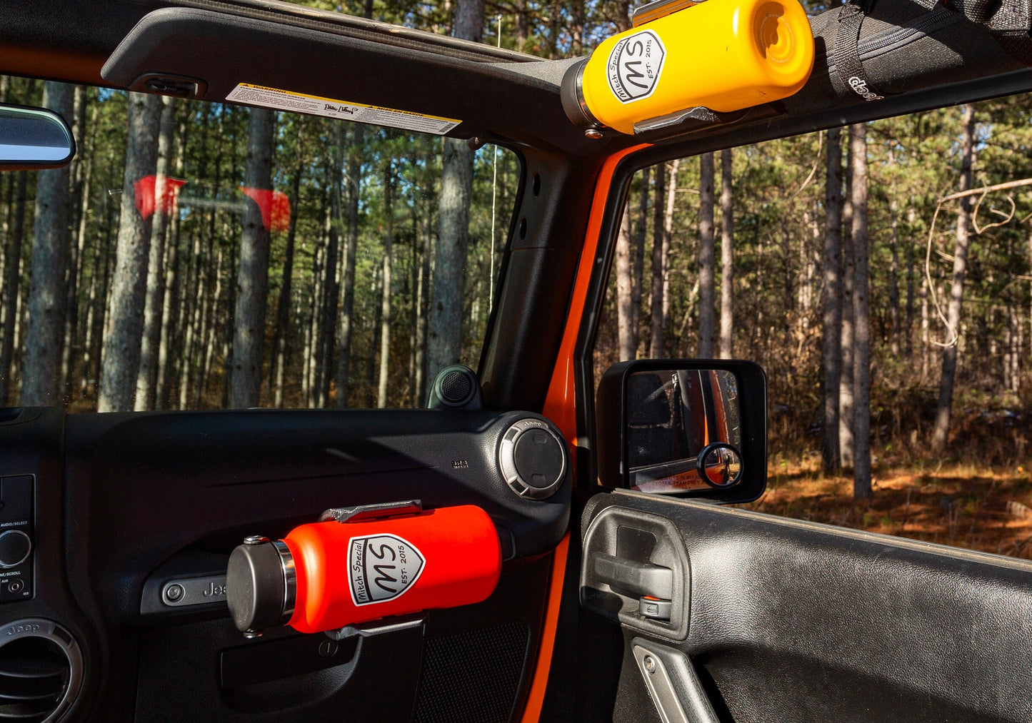 Interior view of a Jeep Wrangler featuring two water bottles, red and yellow, securely mounted on grab handles using custom HD edition water bottle holders, with a scenic forest background highlighting an adventure-ready setup.