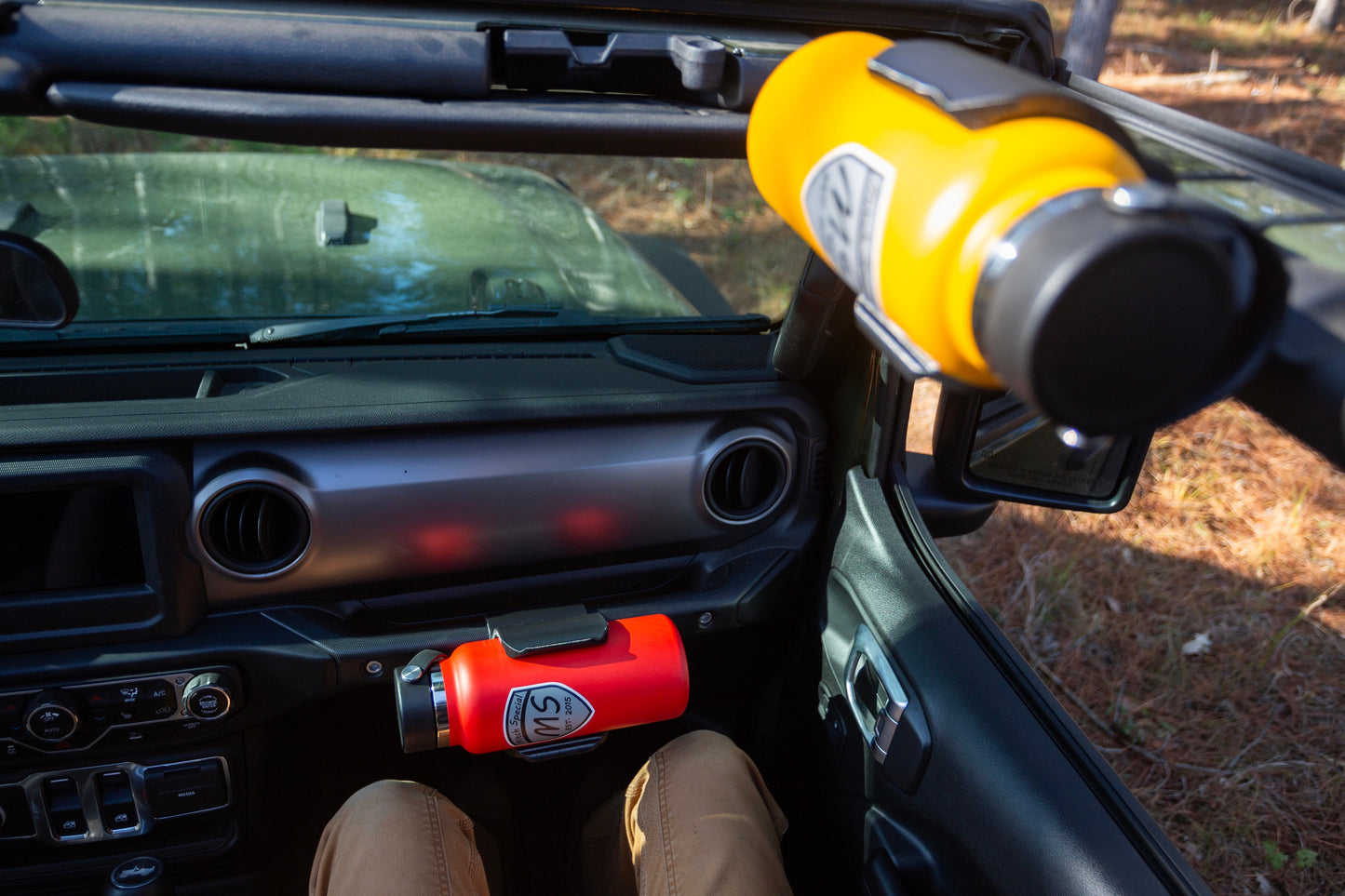 Occupied passenger seat view of Jeep JL interior, featuring Heavy-Duty Water Bottle Holder mounted to grab handle, within easy reach of the passenger, with roll bar water bottle holder also visible, demonstrating the vehicle's versatile storage solutions for outdoor enthusiasts