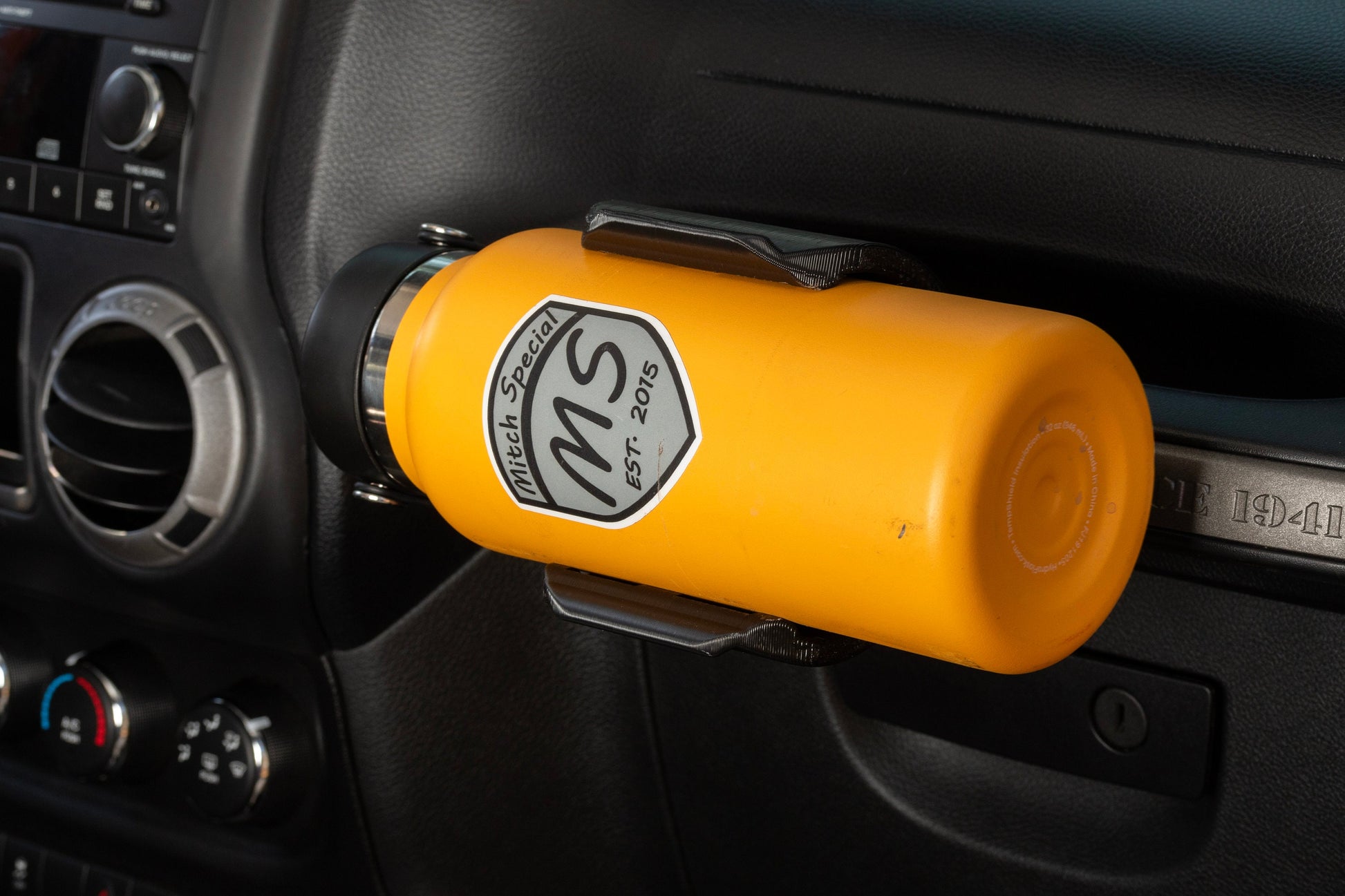 Close-up of a bright yellow water bottle in a holder mounted to the passenger grab handle of a Jeep JK, with a view of the center console at a slight angle, highlighting a functional and stylish accessory for Jeep enthusiasts.