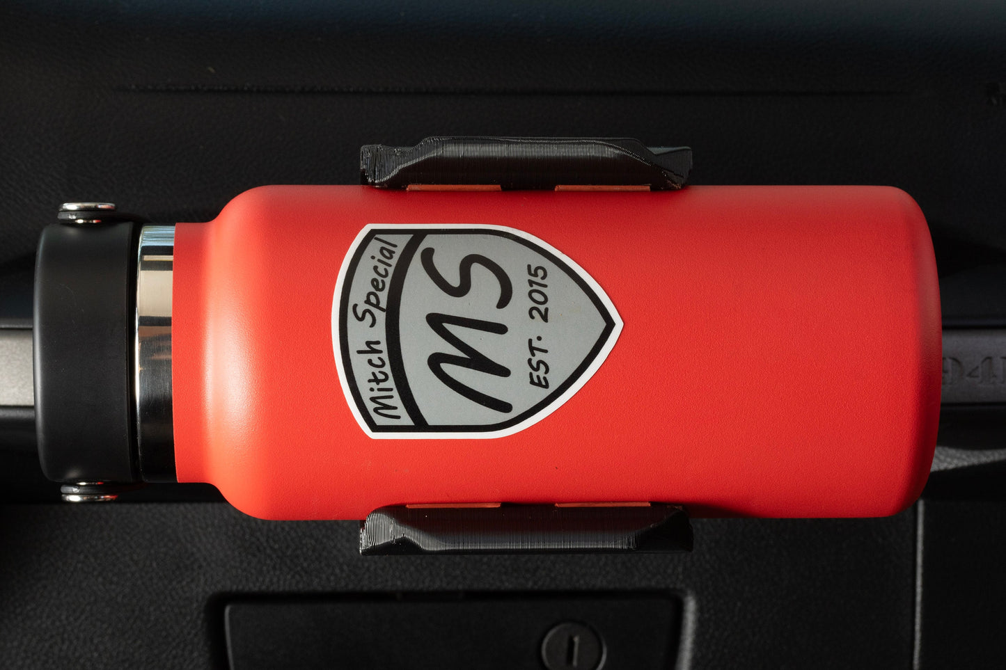 Close-up of a red water bottle labeled 'Mitch Special MS EST. 2015,' secured in a heavy-duty grab handle-mounted holder. Designed for Jeep Wrangler models, this accessory ensures easy access to hydration during off-road trips or daily drives.